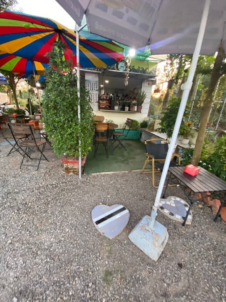 Outdoor coffee kiosk in Cambodia called The Press Day Coffee with colorful umbrellas and wooden furniture surrounded by greenery.